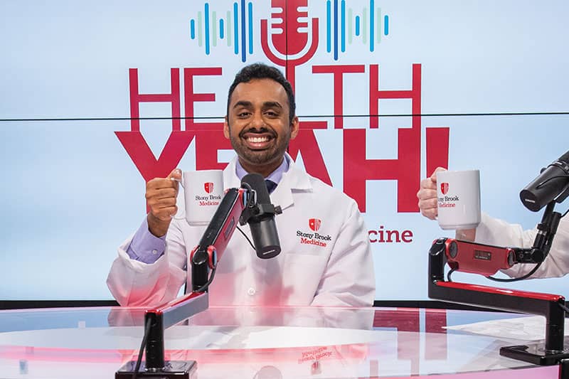 Smiling doctor holding a Stony Brook Medicine mug while recording a podcast titled “Health Yeah!”