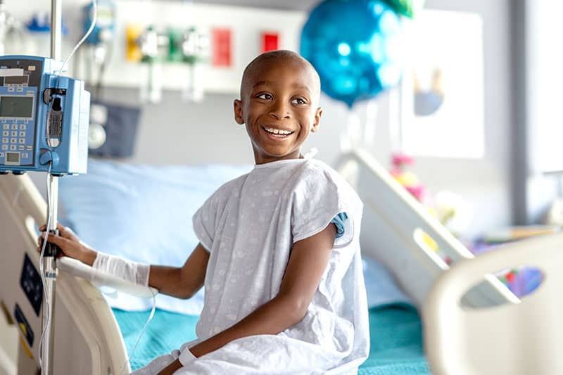 Smiling young boy in a hospital bed wearing a medical gown, receiving IV treatment.