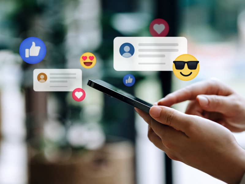Hands holding a smartphone with floating social media icons such as likes, hearts, comments, and emojis